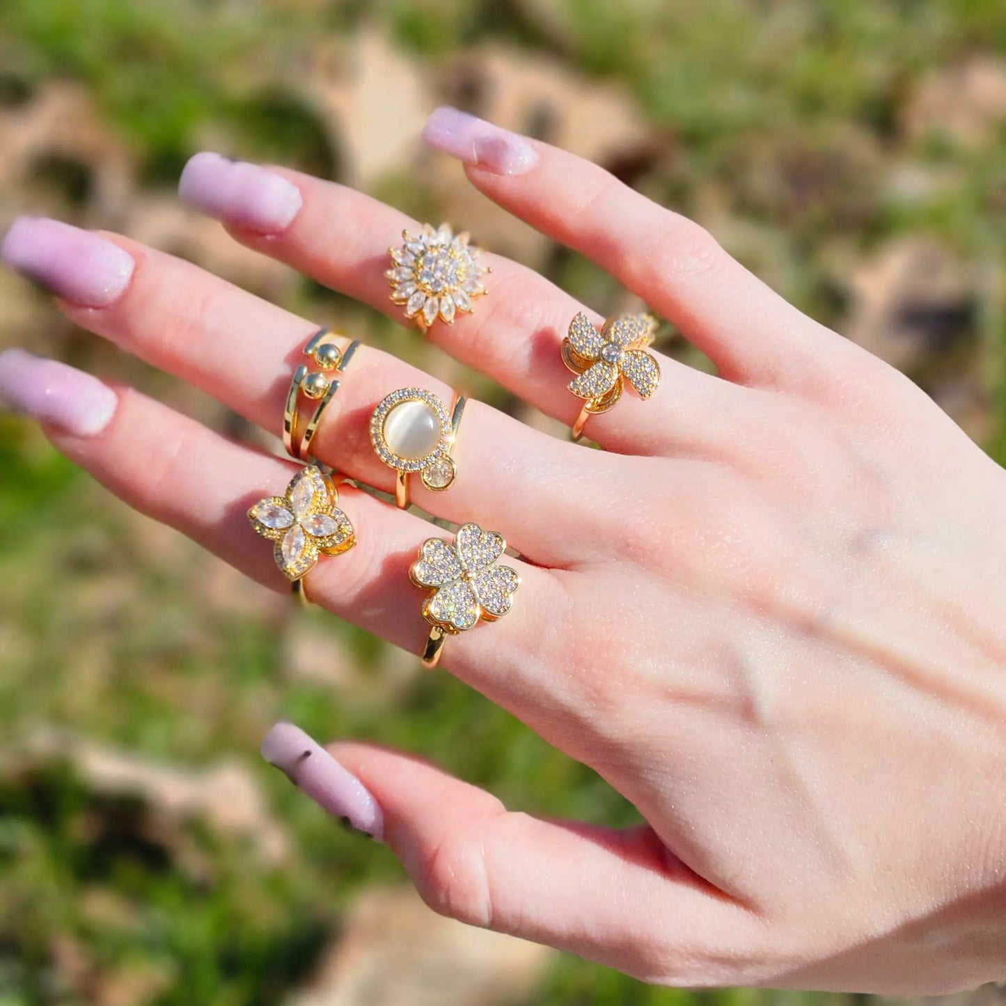 Hand wearing multiple gold rings with floral designs against a blurred natural background