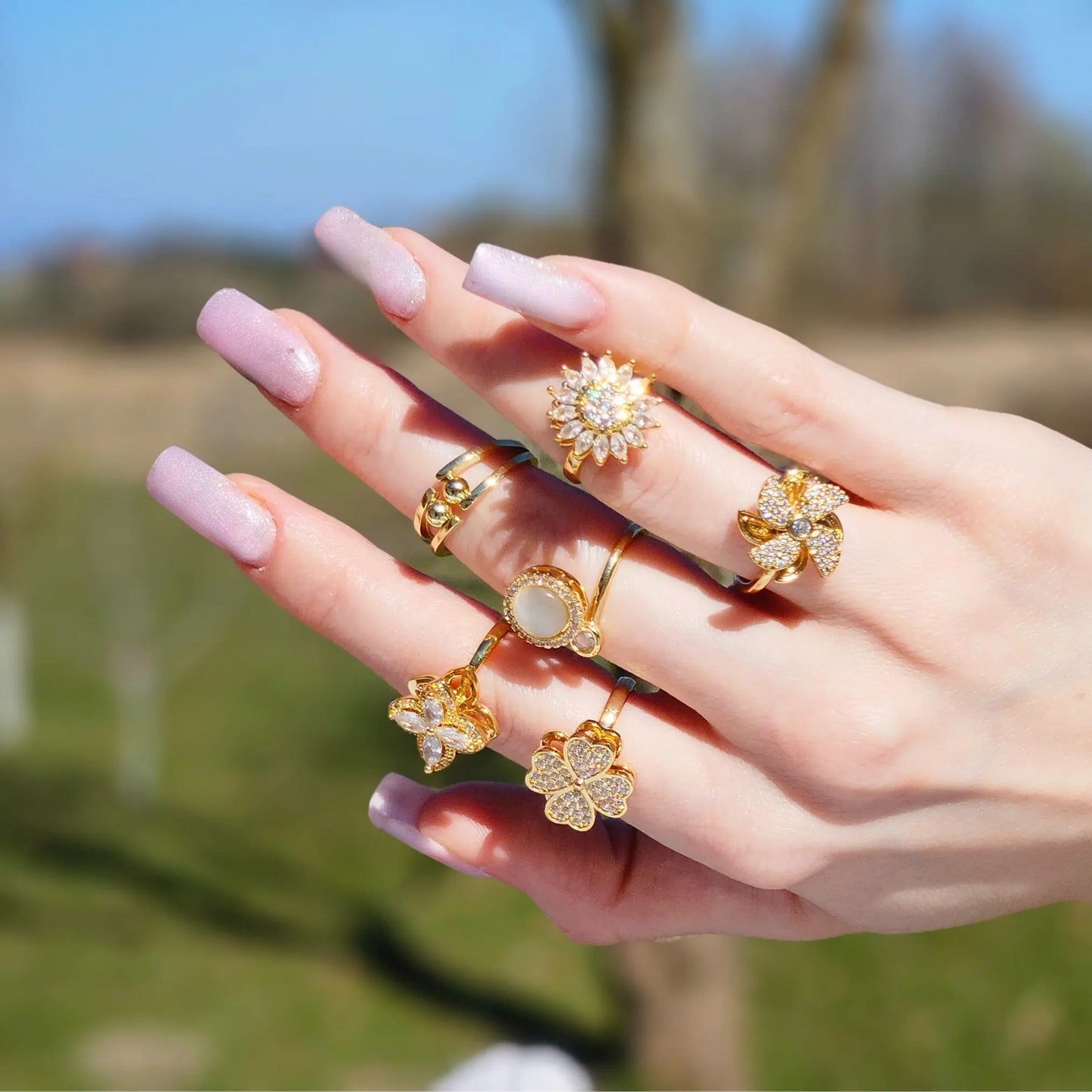 Hand wearing multiple gold rings with floral designs against a blurred natural background